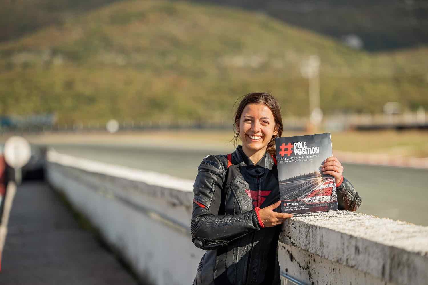 Gina in Lederkombi an der Boxenmauer, im Hintergrund Rennstrecke und Berge, in der Hand der #PolePosition-Ratgeber