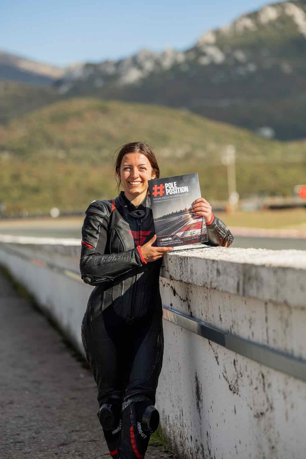 Gina lehnt in Lederkombi an der Boxenmauer und hält den #PolePosition-Ratgeber